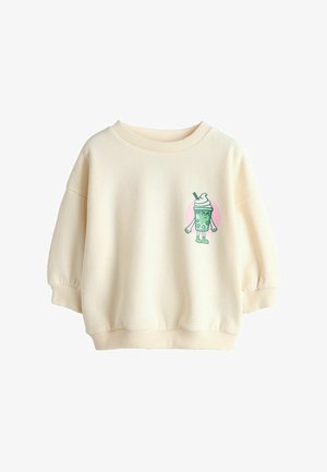 Dolga majica s kremasto barvo in dolgimi rokavi z rebrastimi manšetami ter majhnim risanim likom zelenega mlečnega napitka z rokami in nogami na levi strani prsnega koša.