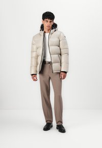 Lichtbeige gewatteerde jas met een rits, voorzien van een capuchon en een gladde, gewatteerde textuur. Draagt over een wit shirt en een bruine broek.