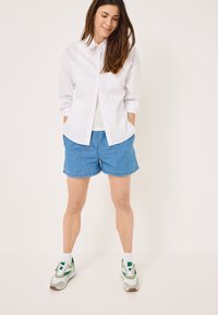 Camicia bianca con bottoni e maniche lunghe, abbinata a shorts di jeans blu. Le calzature includono sneakers grigie e verdi. Outfit semplice e casual.