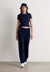 Eine Frau geht drinnen mit einem navyblauen Crop-Top, weit geschnittenen Hosen mit Seitenstreifen, einem Bucket Hat und weißen Sneakers vor einer schlichten weißen Wand.