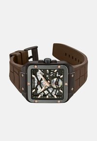 Bruine rubberen horloge met een zwarte vierkante kast, transparante wijzerplaat, gouden accenten en zichtbare bewegingsdetails. Voorzien van een kroon en een textuurband.