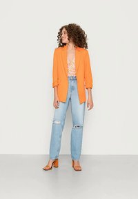 Kvinde med krøllet hår iført en orange blazer, blomstret top, slidte lyseblå jeans og orange højhælede sandaler, stående og kigger til venstre.