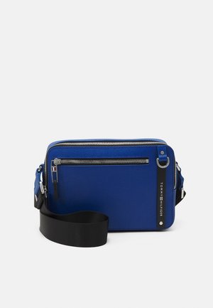 Niebieska prostokątna torba na ramię z przednią kieszenią na suwak, czarnym regulowanym paskiem i metką z logo Tommy Hilfiger po prawej stronie.