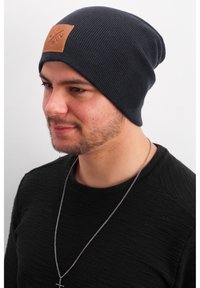Knit blue beanie with a tan leather patch featuring symbols, worn by a person with a textured black sweater and a silver necklace.