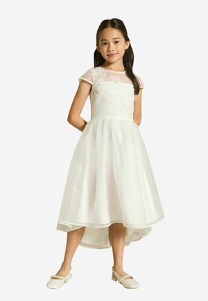 Ragazza giovane in piedi con le mani dietro la schiena, indossa un vestito bianco senza maniche con ricamo floreale e scarpe basse bianche.