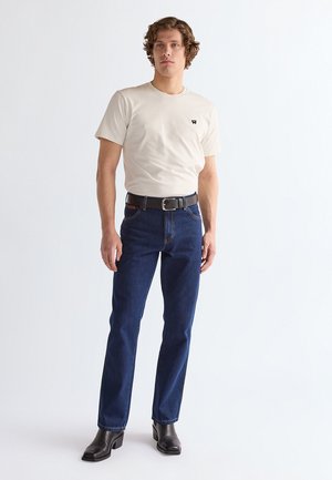 Junger Mann steht, trägt ein beiges kurzärmeliges T-Shirt, dunkelblaue Jeans mit schwarzem Gürtel und schwarze Lederstiefel, vor einfachem Hintergrund.