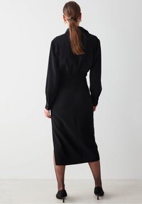 Abito nero a maniche lunghe con vita stretta, colletto alto, spacco posteriore e tessuto morbido. Scarpe con tacco completano l'outfit.
