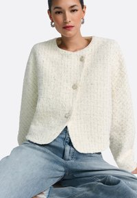 Balta teksturēta jaka ar austa raksta rakstu, trim dekoratīvām pogām un apaļu kakla izgriezumu, kopā ar gaiši ziliem džinsa džinsiem.