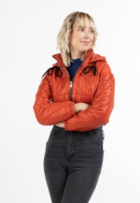 Giacca piumino rossa con finitura lucida, chiusura frontale a zip, cappuccio con cordino e dettagli neri. Abbinata a jeans scuri a vita alta.
