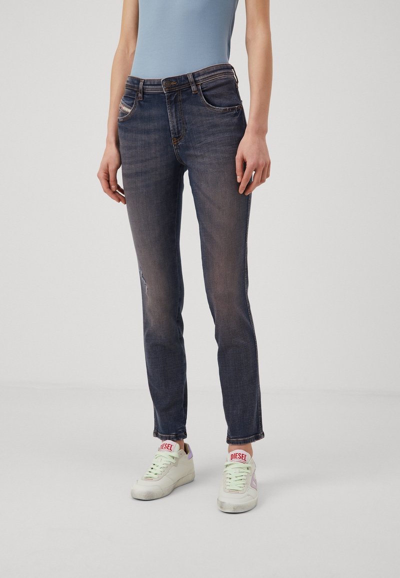 Jean bleu délavé ajusté porté avec un haut sans manches bleu clair et des baskets blanches Diesel avec des détails de logo rouges.