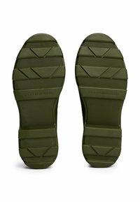 Suelas de zapatos de goma en color verde oliva con surcos y patrones texturizados. Presenta estrías horizontales y diagonales para mayor tracción. La marca es visible.
