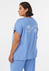 Vaaleansininen puuvillainen t-paita käännetyin hihoin, jossa valkoiset painetut silmät ja teksti takana, suunniteltu mukavuutta ja rentoa istuvuutta varten.