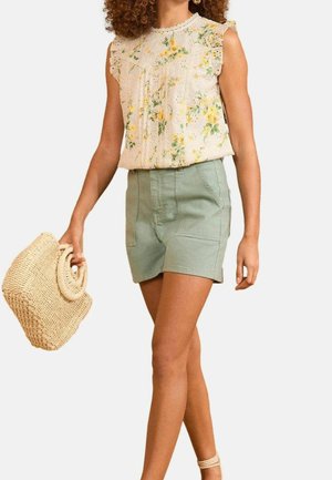 Femme portant un chemisier sans manches à motif floral et un short vert clair, tenant un sac en paille tressée, marchant avec des cheveux bouclés.