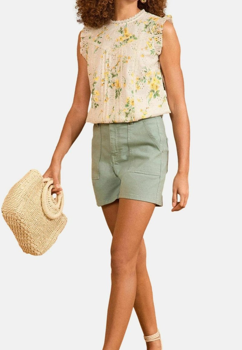 Femme portant un chemisier sans manches à motif floral et un short vert clair, tenant un sac en paille tressée, marchant avec des cheveux bouclés.