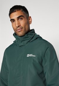 Groene waterdichte jas met een hoge kraag, featuring een logo op de linkerborst. Eenvoudige textuur en lichtgewicht ontwerp voor gebruik buitenshuis.