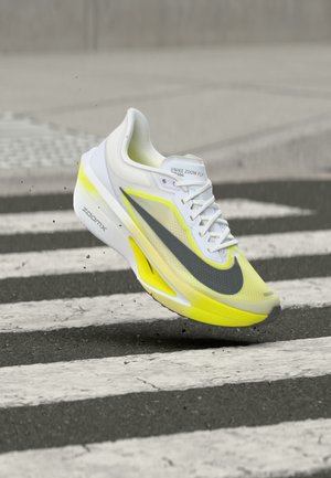 Chaussure de course Nike Zoom Fly blanche et jaune avec le Swoosh gris, en suspension au-dessus d'un passage piéton.