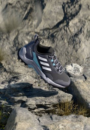 Gray outdoor shoe with mesh upper, blue accents, rubber sole, and three white stripes. Positioned above rocky terrain.