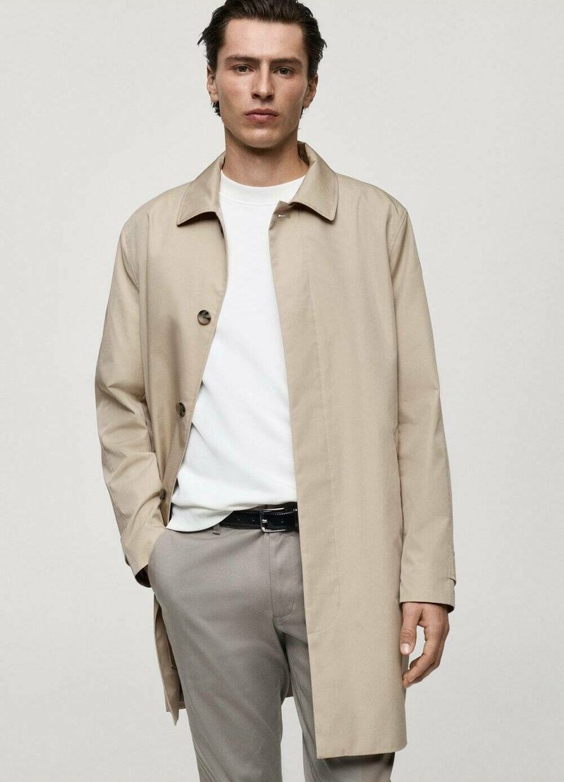 Beige trenchcoat med en klassisk design, med knäppning, spridd krage och långa ärmar, bärs över en vit t-shirt och grå byxor.