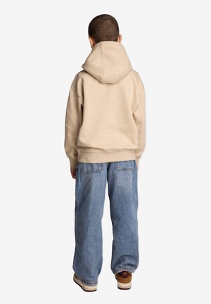Beżowa bluza z dużym kapturem, widziana z tyłu, w połączeniu z luźnymi niebieskimi dżinsami i kremowymi trampkami z czerwonymi akcentami.