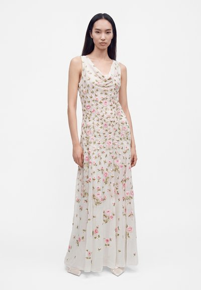 Femme aux longs cheveux foncés portant une robe maxi blanche sans manches avec une broderie florale rose et verte, debout devant un fond blanc.