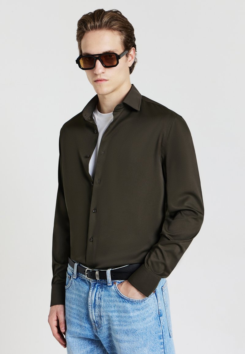Camicia a maniche lunghe verde oliva scuro con colletto button-down, abbinata a una t-shirt bianca e jeans chiari strappati. Cintura nera e occhiali da sole.