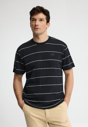 Mężczyzna ubrany w czarną koszulkę z krótkim rękawem z cienkimi białymi poziomymi paskami i beżowe spodnie, stojący z rękami w kieszeniach.