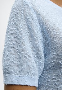 Ανοιχτό μπλε υφασμάτινο πλεκτό με υφή και ανάγλυφο μοτίβο με φουσκάλες σε κοντό μανίκι, που δείχνει τον ώμο και το πάνω μέρος του χεριού.