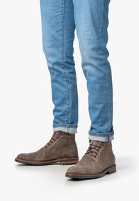 Bottes chelsea en daim marron avec six œillets, lacets marron, et semelle en caoutchouc, associées à un jean tapered bleu clair avec des revers.