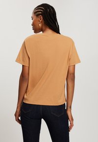 Krótki rękaw, skrócony T-shirt z tanowego bawełnianego materiału. Prosty fason, okrągły dekolt, miękka tekstura, czysta sylwetka, zestawiony z ciemnymi dżinsami.