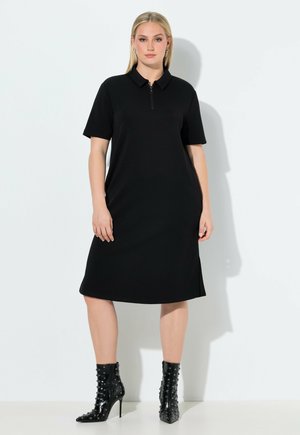 Kvinde iført en sort polo-kjole, der går til knæet, med korte ærmer og sorte støvede ankelstøvler med høje hæle, stående mod en hvid baggrund.