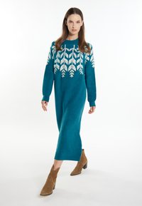 Rochie tricotată cu mâneci lungi, de culoare teal, cu un model alb în formă de frunză pe piept. Materialul pare moale, iar rochia are o croială lejeră până la jumătatea gambei.