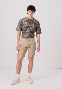 T-shirt camouflage dans des tons terreux avec des manches courtes, assorti à un short cargo beige arrivant aux genoux avec des poches latérales et une coupe droite.