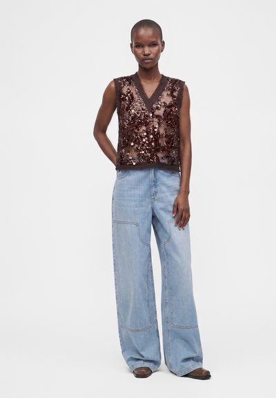 Femme portant un haut sans manches à sequins marron foncé et un jean large bleu clair debout devant un fond blanc uni.