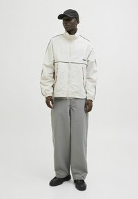 Jachetă albă cu fermoar, cu detalii negre, asociată cu pantaloni largi gri și pantofi negri. Modelul poartă o șapcă neagră. Design simplu, material ușor.