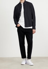 Camicia nera a maniche lunghe sopra una maglietta bianca a collo rotondo, abbinata a jeans scuri e sneakers bianche. Tessuto morbido, vestibilità rilassata.
