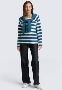 Długi rękaw w paski w kolorach turkusowym i białym, z dzianinowym swetrem związanym wokół szyi. Połączony z ciemnymi jeansami i białymi tenisówkami.