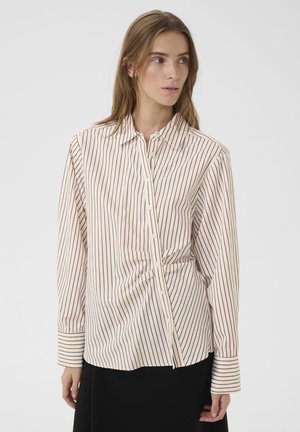 Kvinde iført en hvid bluse med lodrette brune striber og et sort skørt, der kigger mod sin højre side mod en ensfarvet baggrund.