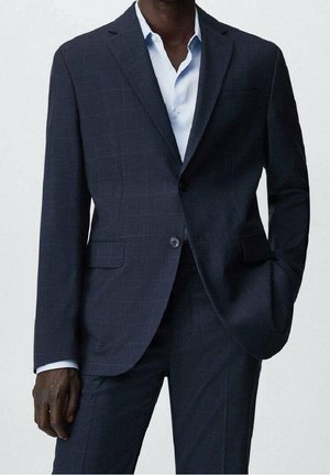 Hombre vestido con un traje azul marino oscuro y una camisa de vestir azul claro, con una mano en el bolsillo, de pie frente a un fondo claro y liso.