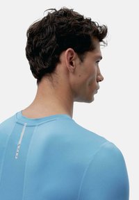 Šviesiai mėlyna sportinė marškinė su apvaliu iškirpimu ir subtilia tekstūra; gale yra vertikali balta detalė.