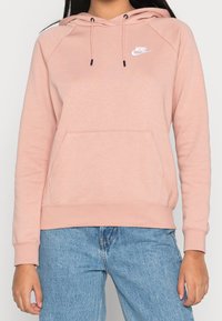 Lichtroze Nike-hoodie met koordjes, kangoeroezak en geribbelde manchetten. Voorzien van een klein wit logo op de bovenkant van de borst. Zacht materiaal.