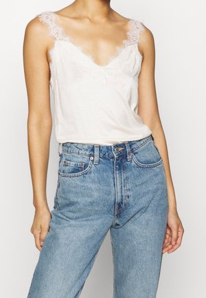 Kobieta ubrana w biały jedwabny top wykończony koronką, wpuszczony w jasnoniebieskie, wysokie dżinsy, stojąca na jasnym tle.