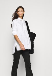 Camisa branca de botões com bolinhas pretas, mangas enroladas, fendas laterais, combinada com calças pretas largas e uma bolsa preta texturizada.