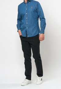 Niebieska koszula dżinsowa z kołnierzykiem zapinanym na guziki, długimi rękawami i małym haftowanym logo. Połączona z czarnymi spodniami i białymi trampkami.