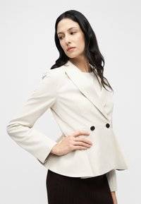 Blazer beige sur mesure avec un ourlet à basque, doté de deux boutons noirs, de manches longues et d'un tissu lisse et structuré.