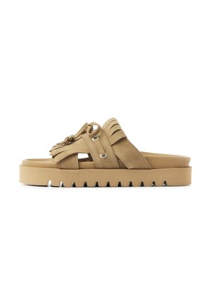 Beige suede slide sandal with thick ridged sole, fringe details, and metallic eyelets on crossed straps.
