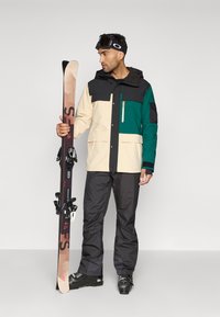 Casaco de esqui em bege, verde e preto; bolsos frontais; combinado com calças de esqui pretas e esquis. Botas de esqui pretas completam o visual.