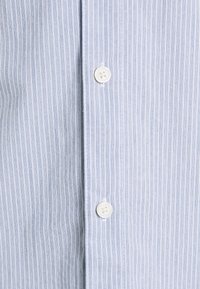 Tela de camisa de rayas azul claro con finas líneas verticales, con botones blancos y una textura suave. Sin patrones o acentos adicionales.