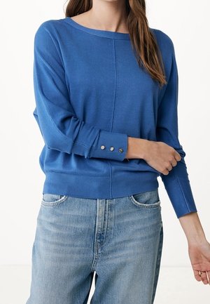 Kobieta ubrana w niebieski sweter z długim rękawem z trzema guzikami na mankiecie, w połączeniu z jasnymi dżinsami o jasnym odcieniu.