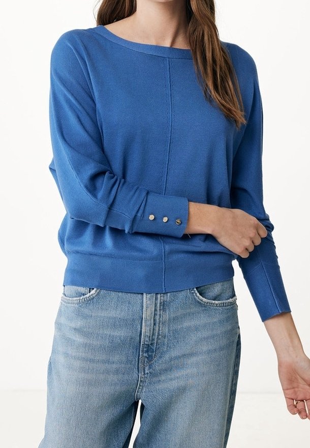 Femme portant un pull à manches longues bleu avec trois boutons sur le poignet, associé à un jean en denim délavé clair.