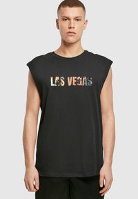Melnā bezdarbības T-krekla, uz kura attēlots grafisks prints ar uzrakstu "LAS VEGAS" daudzkrāsainās burtos, kā arī attēli ar palmām un ēkām.